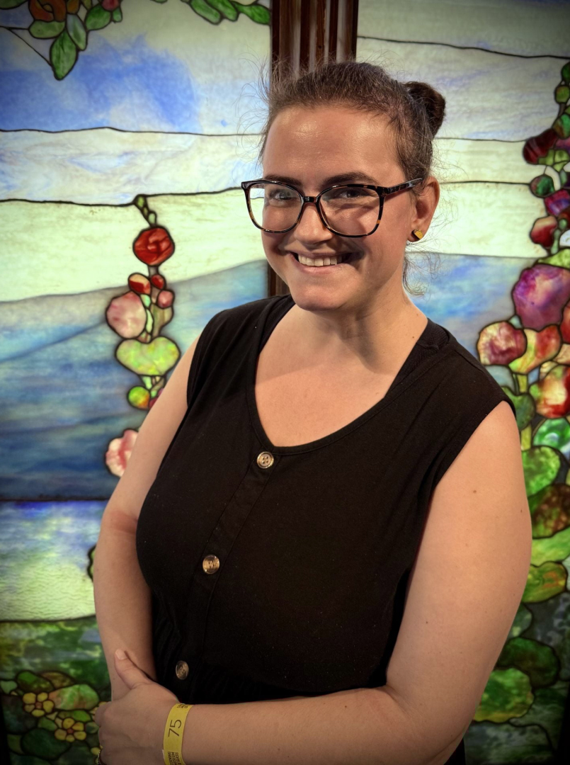 A young person with glasses and a black shirt in front of a stained glass window.